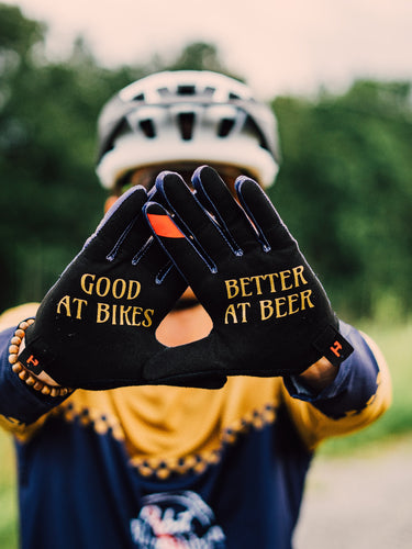 Person wearing gloves with 'Good at Bikes, Better at Beer' text on a blurred natural background