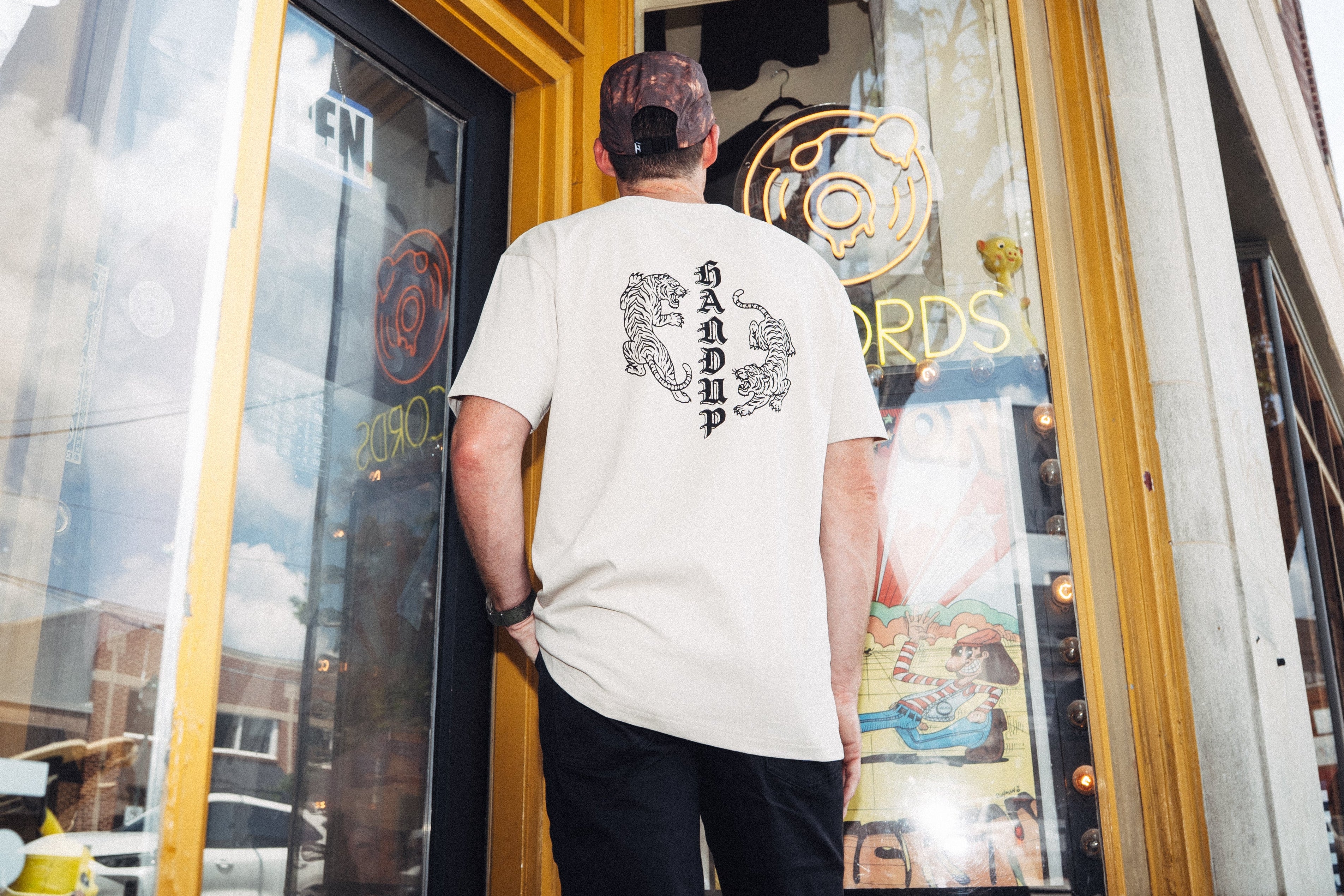 A man stands with his back to the camera, wearing a cream-colored t-shirt with a black graphic print on the back, looking out of a storefront window. The shirt features a large black graphic of a figure with a sword on the back.