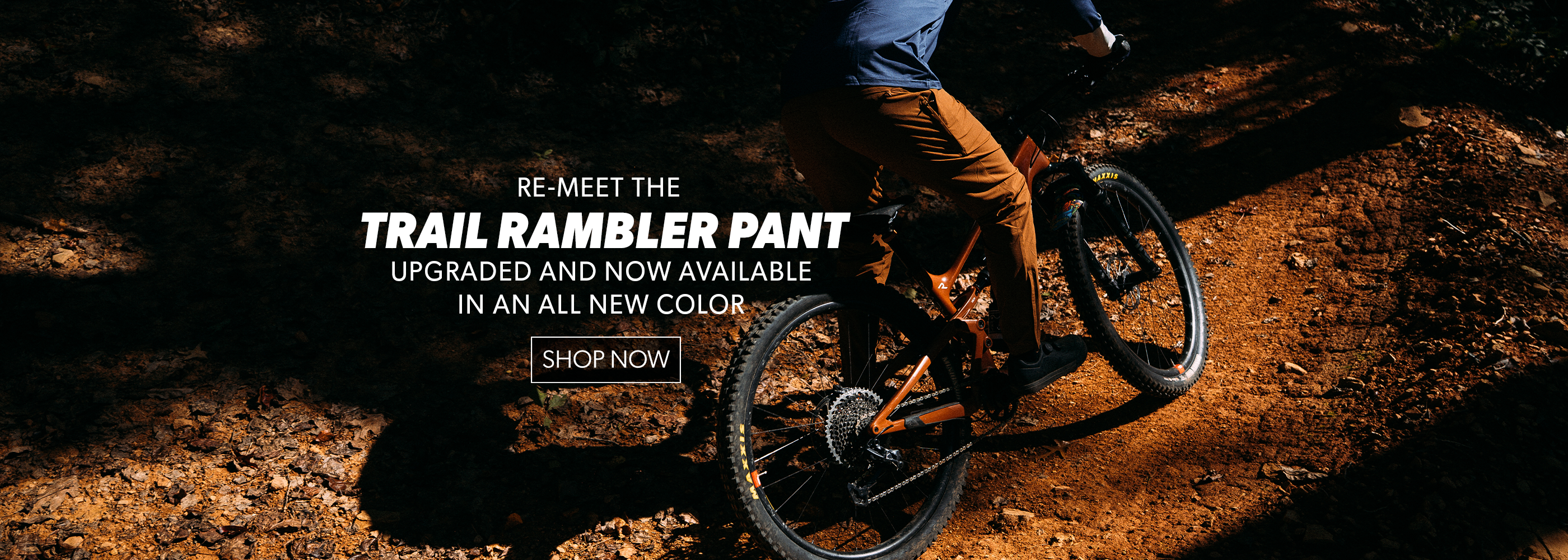 Person riding a mountain bike on a trail with promotional text for Trail Rambler Pant.