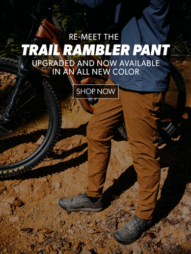 Person wearing brown trail rambler pants standing next to a bicycle on a dirt path.