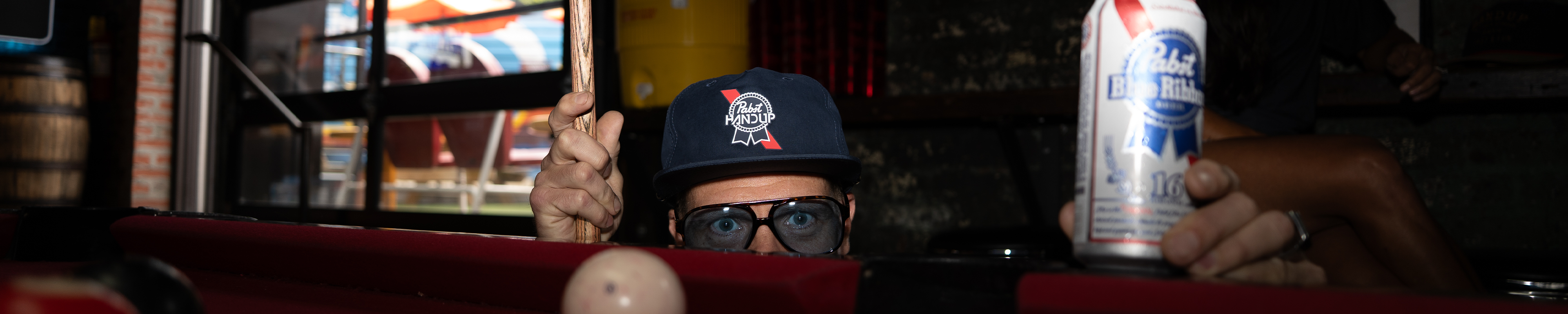 a person with a hat and sunglasses peaking over the edge of a pool table holding a beer