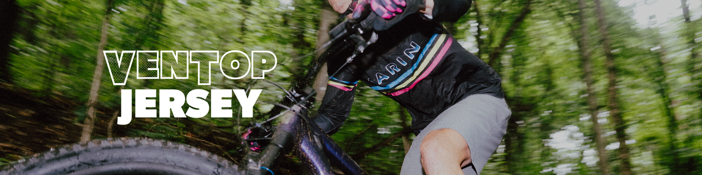 A mountain biker riding through a blurred forest with the text 'VENTOP JERSEY' overlaid on the left side. The rider is wearing a black jersey with pink, blue, and white stripes, gray shorts, and gloves.