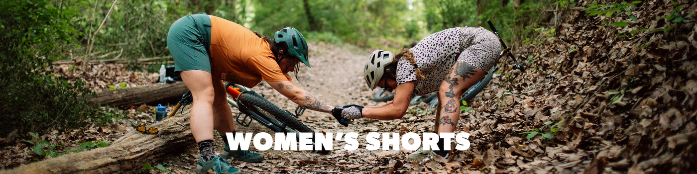 Two people in mountain biking gear working on a log in a forest with 'Women's Shorts' text overlay.