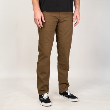 A person wearing brown straight-leg work pants, a black t-shirt, and black Vans sneakers, standing against a light gray studio background.