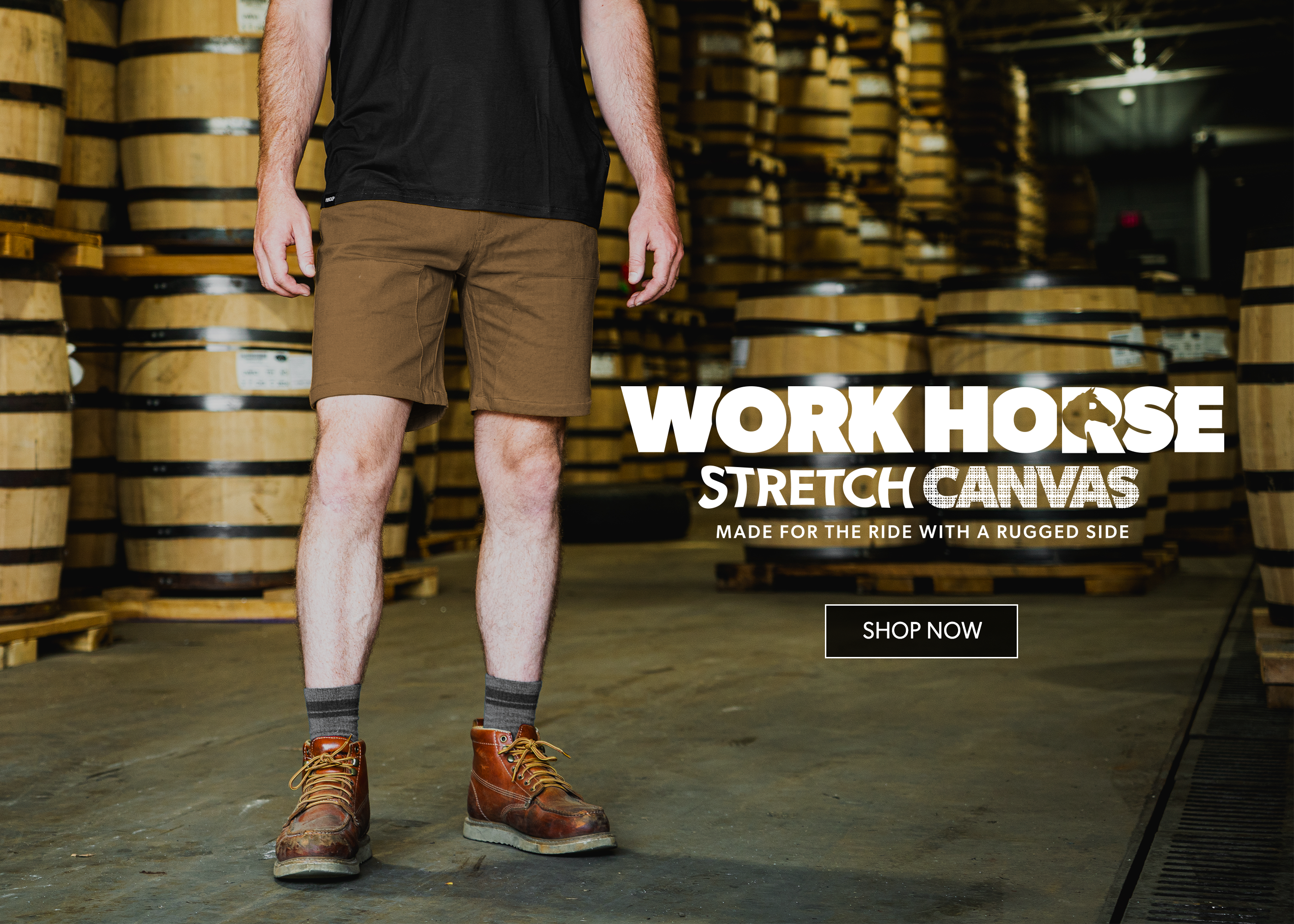A person standing in a warehouse with stacked wooden barrels, wearing brown canvas work shorts, a black t-shirt, gray socks, and brown leather work boots. Large text on the right reads 'WORK HORSE STRETCH CANVAS' with a 'SHOP NOW' button below.
