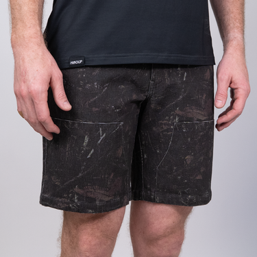 A person wearing dark charcoal shorts with a distressed, scuffed texture and a black t-shirt, shown from the chest down against a light gray studio background.