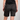 Rear view of a person wearing black camouflage shorts with a subtle bark pattern, paired with a black t-shirt, against a light gray studio background.