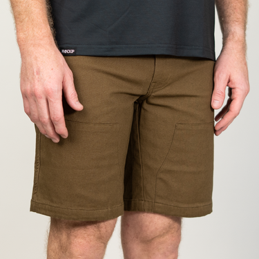 A close-up of a person wearing olive brown cargo shorts and a dark charcoal t-shirt. The shorts feature side pockets and a clean, straight-leg fit, photographed against a neutral light gray background.
