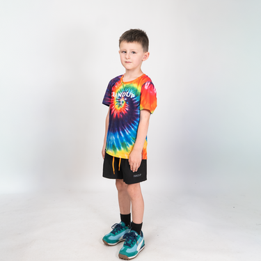 A young boy stands against a light gray studio background wearing a vibrant tie-dye t-shirt with a spiral pattern in blue, yellow, orange, and red, featuring the brand name 'KANDOR' on the chest. He is also wearing black shorts and teal sneakers.