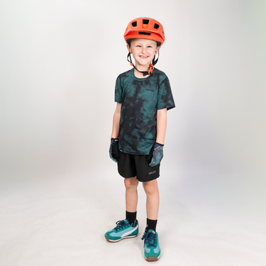 A young boy wearing a dark green and black tie-dye short-sleeve jersey, black shorts, teal and green mountain biking shoes, a bright orange helmet, and black gloves, standing against a light gray studio background.
