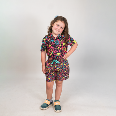 A young girl modeling a matching short-sleeve collared shirt and shorts set with a colorful abstract pattern, standing against a light gray studio background.