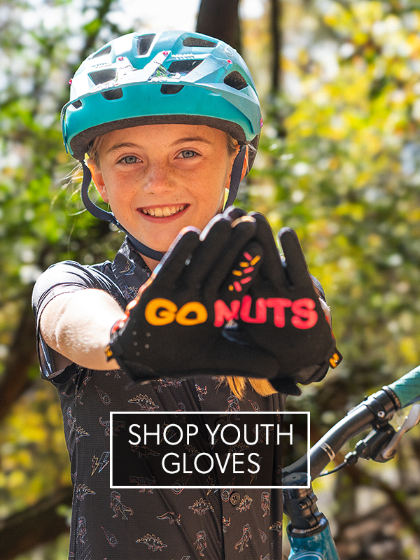 Child wearing a helmet and gloves with 'GO NUTS' branding, holding out a glove towards the camera with a blurred natural background.